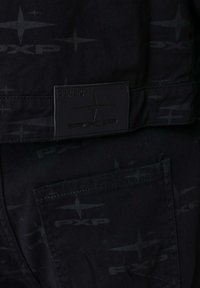 Pantalones de lona negros con un patrón de logotipo tono sobre tono y una etiqueta rectangular de la marca cosida en la cintura, con un bolsillo trasero visible.