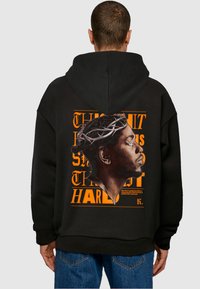 Sort oversized hoodie med et stort, farverigt grafik på ryggen af en profil, omkranset af orange tekst med forskellige skrifttyper.
