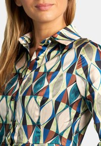 Camisa de botones con estampado que presenta un diseño geométrico en verde, azul, marrón y negro, hecha de una tela suave con un cuello clásico.