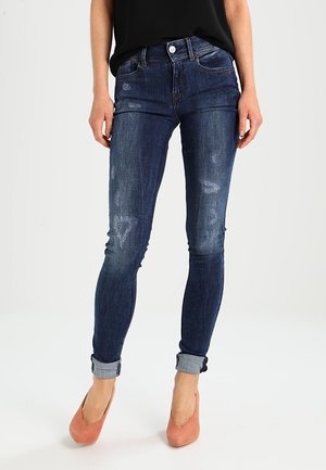 Jean skinny en denim bleu foncé avec un design délavé, plusieurs accents usés et des ourlets roulés, associés à des chaussures de couleur pêche.