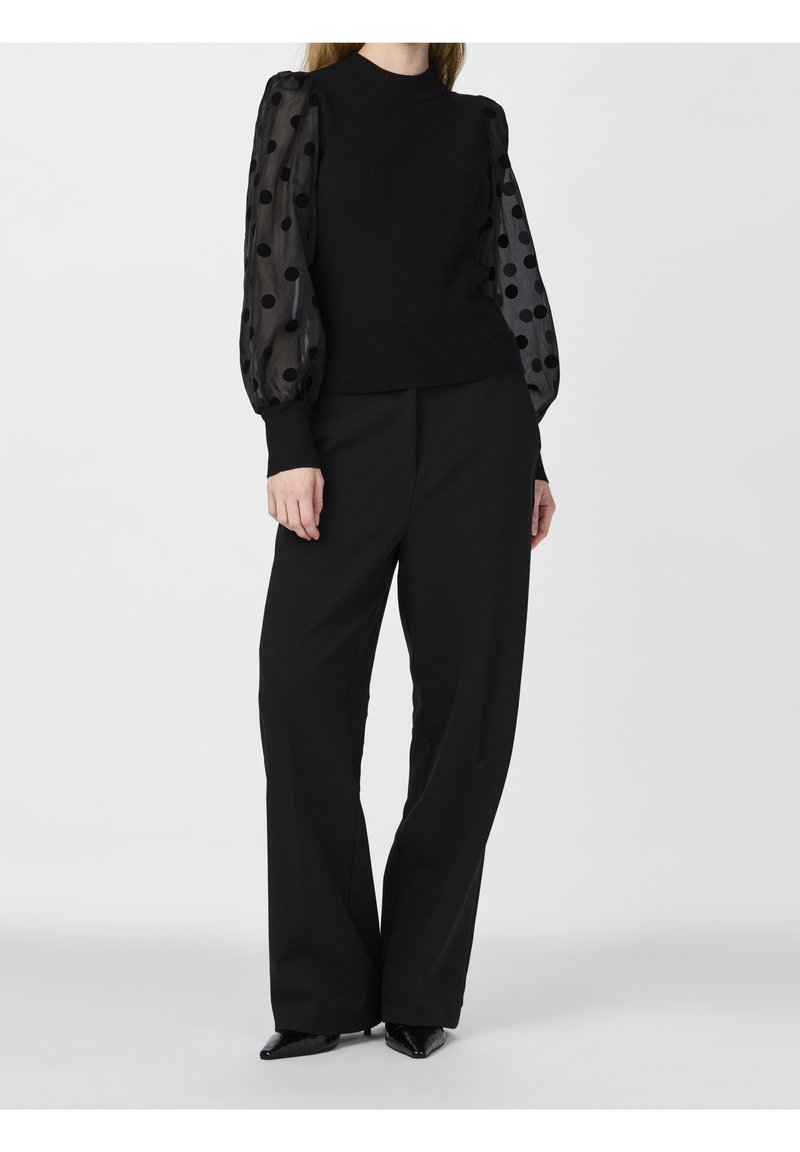 Femme portant un haut noir à col montant avec des manches bouffantes transparentes à pois et un pantalon noir à jambes larges sur un fond uni.