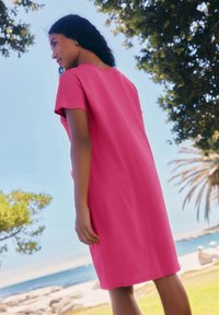 Robe en coton rose à manches courtes, coupe décontractée, longueur genou, texture lisse, avec un col rond. En arrière-plan, des arbres et une vue sur l'océan.