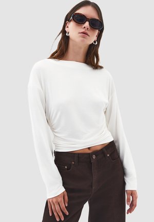 Blusa blanca de manga larga con escote barco, detalle anudado en la cintura, combinada con pantalones de tiro alto marrón oscuro, textura de tela suave.
