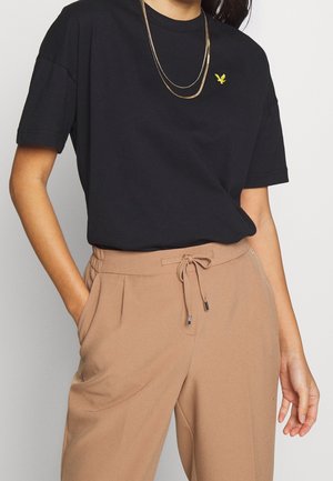 Osoba ubrana w czarną koszulkę z krótkim rękawem z małym żółtym logo ptaka i warstwowe złote naszyjniki, w połączeniu z wysokim stanem beżowych spodni ze sznurkiem.