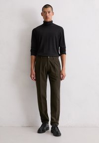Zwarte coltrui, losse pasvorm, gecombineerd met olijfgroene geplooide broek. Zwarte veterschoenen maken de outfit af. Eenvoudige achtergrond.
