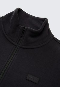 Schwarze Zip-Jacke aus Fleece mit Stehkragen, glatter Textur und einem kleinen schwarzen Markenlabel auf der Brust.