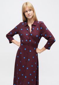 Bordó ruha kék pöttyökkel, hosszú ujjú, gombos elejű és galléros. Sima anyagból, enyhén bővebb fazonú, térd alá érő hosszúságú.