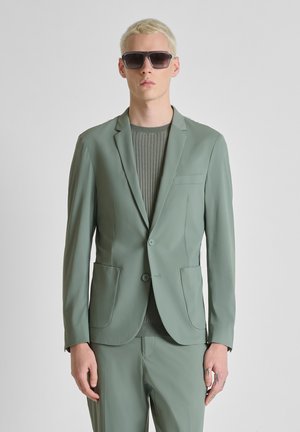 Persona con cabello rubio corto, gafas de sol oscuras, traje verde claro y suéter gris acanalado sobre un fondo liso.