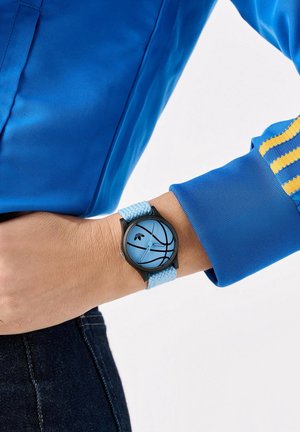 Poignet portant une montre bleue avec un cadran à motif basket-ball et un bracelet en tissu bleu clair, associée à une veste bleue et un jean foncé.