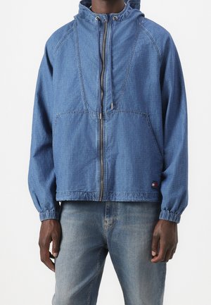 Mężczyzna w niebieskiej kurtce z kapturem zapinanej na zamek, z elastycznymi mankietami i przednimi kieszeniami, w połączeniu z jasnoniebieskimi dżinsami.