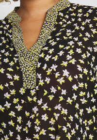 Blouse noire avec un motif floral de fleurs blanches et jaunes. Le col est texturé avec un motif pied-de-poule et une patte de boutonnage sans boutons.