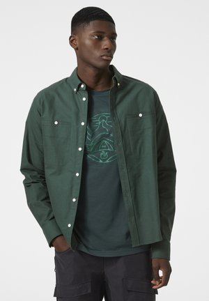 Groene button-up shirt met twee borstzakken, gedragen over een donkergroen graphic t-shirt. Gladde textuur, casual ontwerp, strakke pasvorm.