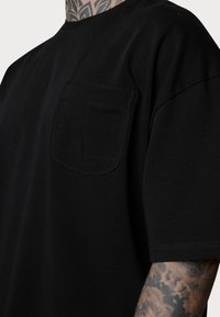 T-shirt en coton noir avec col rond et manches courtes, doté d'une poche cousue sur le côté gauche de la poitrine et d'une texture lisse.