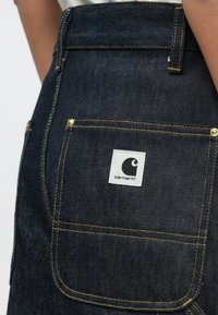 Temno modri žep na kavbojkah iz jeansa z rumenim šivom in belo oznako znamke Carhartt, ki jo nosi oseba v belem vrhu.