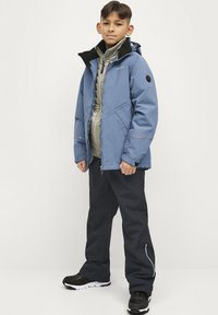 Blauwe waterdichte jas met zwarte accenten, gelaagd over een lichtgrijze gewatteerde binnenvest. Draag het met donkere broek en zwarte sneakers.