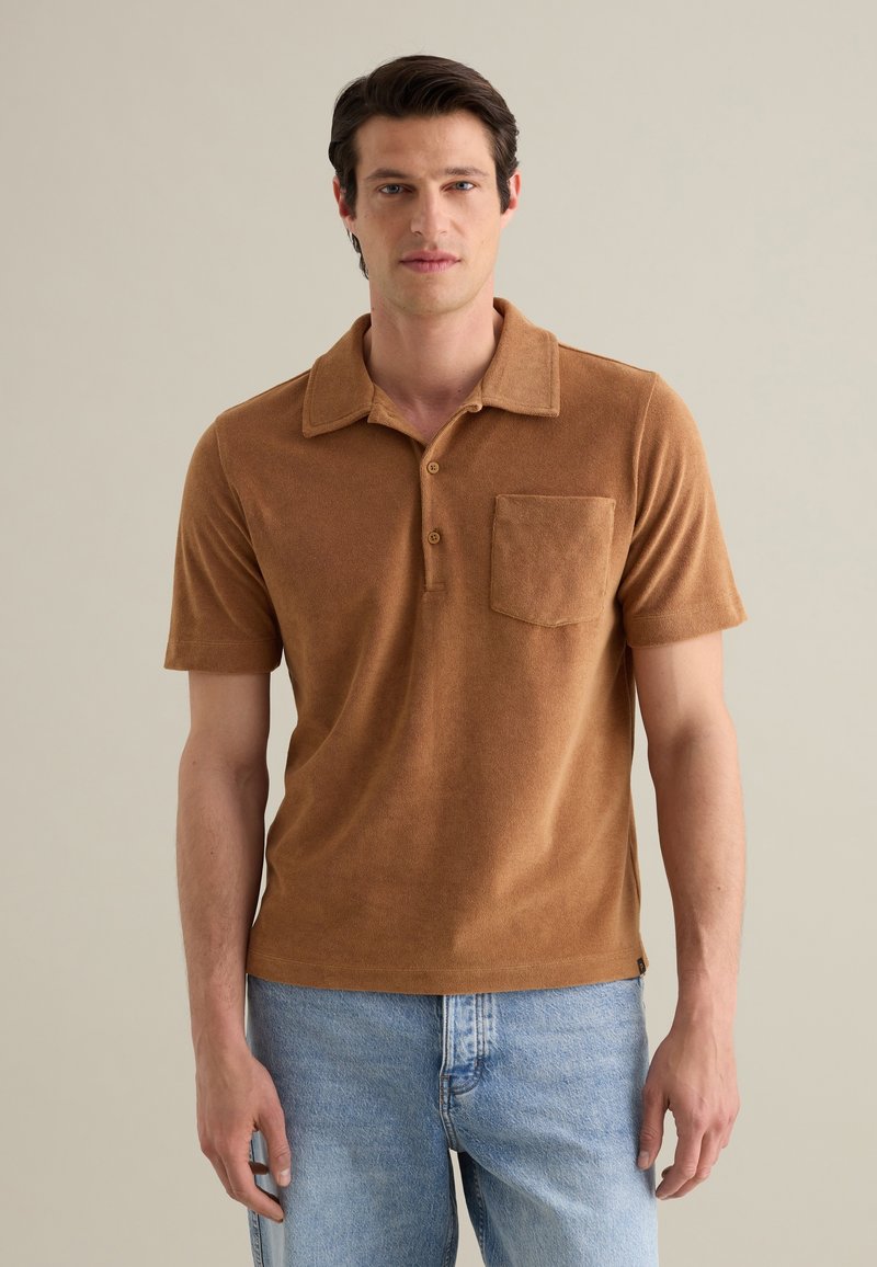 Polo brun en tissu doux, avec un col, trois boutons et une poche poitrine. Porté avec un jean bleu clair.