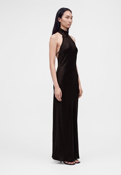 Femme aux longs cheveux noirs, portant une robe noire en satin sans manches à col halter et des talons noirs à lanières, debout devant un fond clair uni.