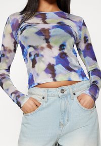 Långärmad topp i ett lätta, genomskinligt tyg med ett abstrakt mönster i blått, grönt och brunt, parat med ljusblå denimshorts.