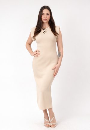 Mujer de pie con un vestido midi sin mangas beige con hombreras acolchadas, lleva tacones abiertos y un collar grande, posando con una mano en la cadera.