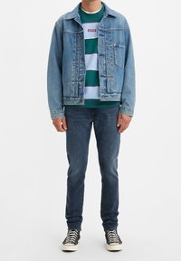 Hellblaue Jeansjacke, türkisfarbener gestreifter Pullover, dunkle Jeans und schwarze Sneakers mit weißen Sohlen. Das Model steht vor einem einfarbigen Hintergrund.