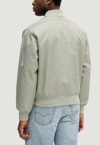 Giacca bomber grigia in tessuto liscio e leggero; presenta un colletto e polsini a costine, tasca con zip sulla manica e vita elastica.