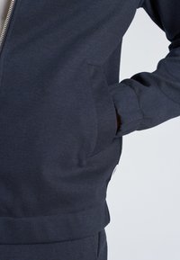 Main dans la poche d'une veste zippée bleu marine foncé avec poignets et taille côtelés sur un fond clair uni.