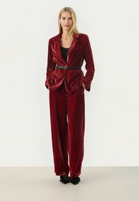 Completo in velluto rosso con giacca aderente, pantaloni larghi e cintura nera strutturata. Tessuto morbido, maniche a sbuffo e design a gamba larga.