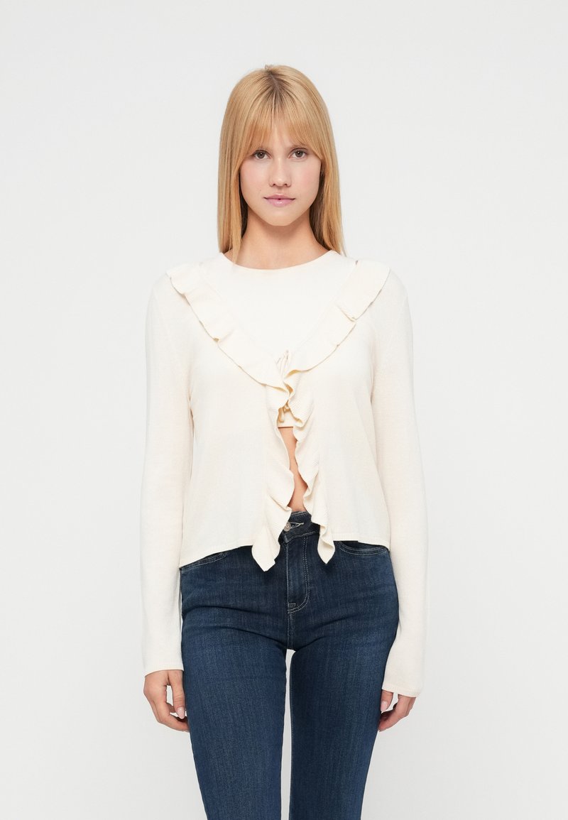 Κρεμ ζακέτα με μακριά μανίκια, διακοσμημένη με βολάν στο μπροστινό μέρος και κλείσιμο με κορδόνι. Συνδυάζεται με σκούρο μπλε εφαρμοστά τζιν. Λεία υφή.