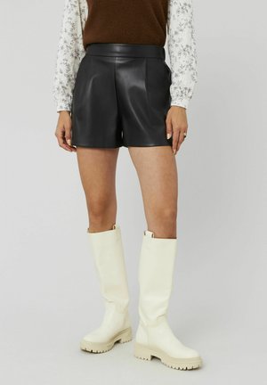 Shorts en cuir noir taille haute avec des plis à l'avant, associés à des bottes en caoutchouc blanches montantes avec une semelle épaisse.