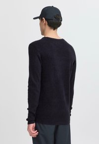Pulover tricotat de culoare bleumarin, cu mâneci lungi, gât rotund și mansete cu model din bumbac. Textură moale, croială lejeră și cusături minime. Purtați cu o șapcă închisă la culoare.