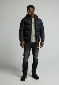 Schwarze Daunenjacke mit Kapuze, mit Reißverschlusstaschen, kombiniert mit einem hellgrünen Hemd und ausgefransten schwarzen Jeans. Schwarze Schuhe runden das Outfit ab.