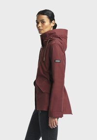 Veste imperméable bordeaux avec capuche, cordons de serrage ajustables, fermeture éclair, poches latérales et un logo sur la manche. Texture de tissu lisse.