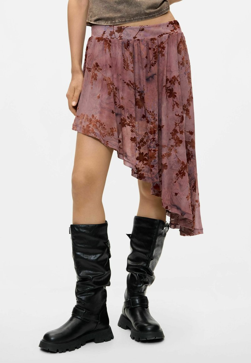 Woman wearing an asymmetrical dusty pink floral skirt and black knee-high leather boots standing against a light background.