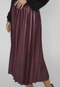 Mujer que lleva una falda midi plisada de cintura alta en burdeos y una camiseta de manga larga negra y suave, con las manos descansando cerca de la cintura.