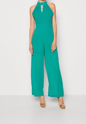 Türkiser Jumpsuit mit Halterneck, Schlüssel-Ausschnitt, weitem Bein und fließendem Stoff; kombiniert mit goldenen Sandalen mit Absatz.