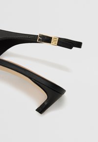Black leather high-heeled sandal with a thin ankle strap and gold-tone buckle. Features a textured design and a subtle curved heel.