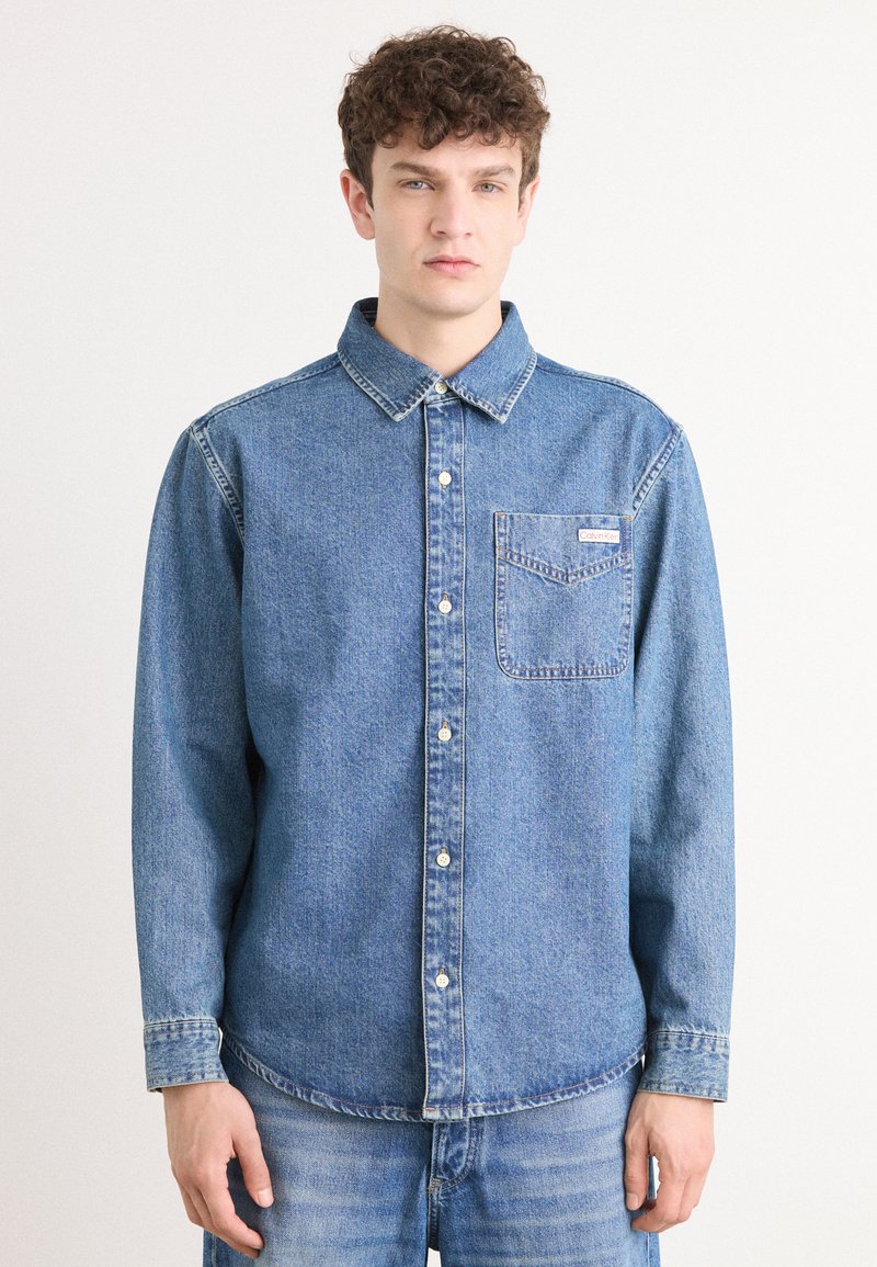 Camisa de mezclilla de botones, azul claro, con un bolsillo en el pecho y una etiqueta de marca. Presenta un cuello clásico y mangas largas con puños de botón.