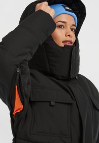 Sort polstret jakke med høj krave, lynlåslomme i siden og orange meshfor. Har en robust lukning og slidstærkt stof.