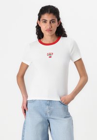 White ribbed t-shirt with a red round neckline, featuring an embroidered logo and cherry design on the chest, paired with light blue jeans.