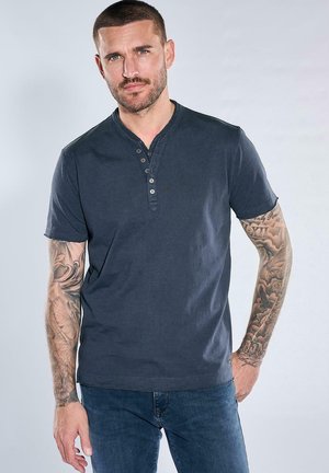 Mann mit kurzen Haaren und Ärmeltätowierungen trägt ein dunkelblaues Henley-Shirt und Jeans, steht mit einer Hand in der Tasche vor einem einfarbigen Hintergrund.