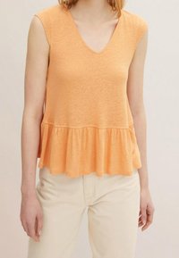 Sleeveless light orange V-neck top with ruffled hem worn with off-white high-waisted pants on a standing person.