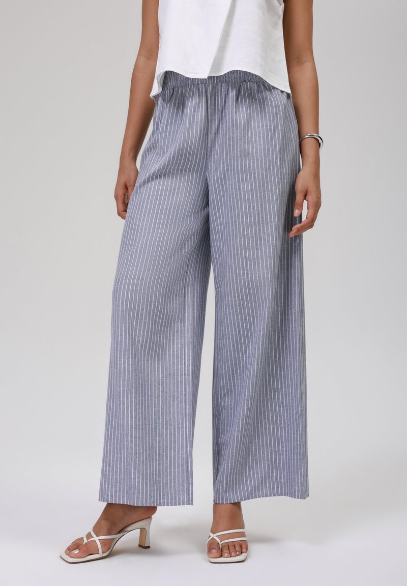 Femme portant un pantalon large à rayures verticales bleu et blanc, un haut sans manches blanc et des sandales à talons blanches, sur un fond gris clair.