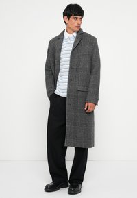 Gray plaid overcoat with long sleeves, a notched collar, and side pockets, layered over a light striped polo shirt and black trousers.