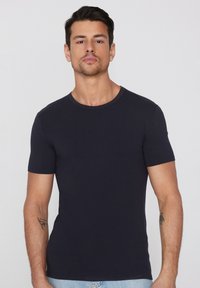 Mężczyzna o krótkich ciemnych włosach i jasnym zarostem nosi czarną jednolitą koszulkę z krótkim rękawem oraz jasnoniebieskie dżinsy, stojąc na białym tle.