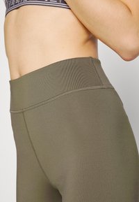 Leggings verde oliva a vita alta con una superficie liscia e dettagli cuciti lungo le cuciture. Una vista ravvicinata mette in risalto la vestibilità del tessuto.