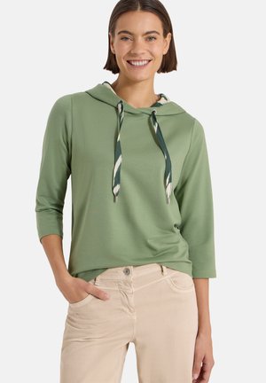 Femme portant un sweat à capuche vert clair avec des cordons à motifs et un pantalon beige, debout avec une main dans la poche, souriant à la caméra.