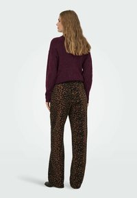 Leopardprintbukser i brun og sort, parret med en mørke lilla, strikket sweater. Model vender væk, hvilket viser den afslappede pasform og tekstur.