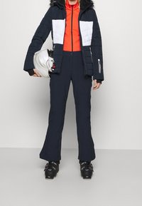 Giacca da sci imbottita nera con pannelli bianchi, maglione arancione con cerniera, pantaloni da sci blu navy. Scarponi da sci neri. Casco tenuto nella mano sinistra.