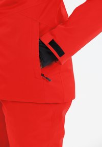 Rode waterdichte jas met zwarte accenten, voorzien van een ritssluiting aan de zijkant en klittenbandsluitingen bij de manchetten voor verstelbaarheid.