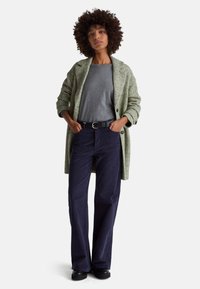 Zielony płaszcz w jodełkę na szarej koszuli, w połączeniu z granatowymi sztruksowymi spodniami i czarnym paskiem. Całość dopełniają gładkie czarne buty.